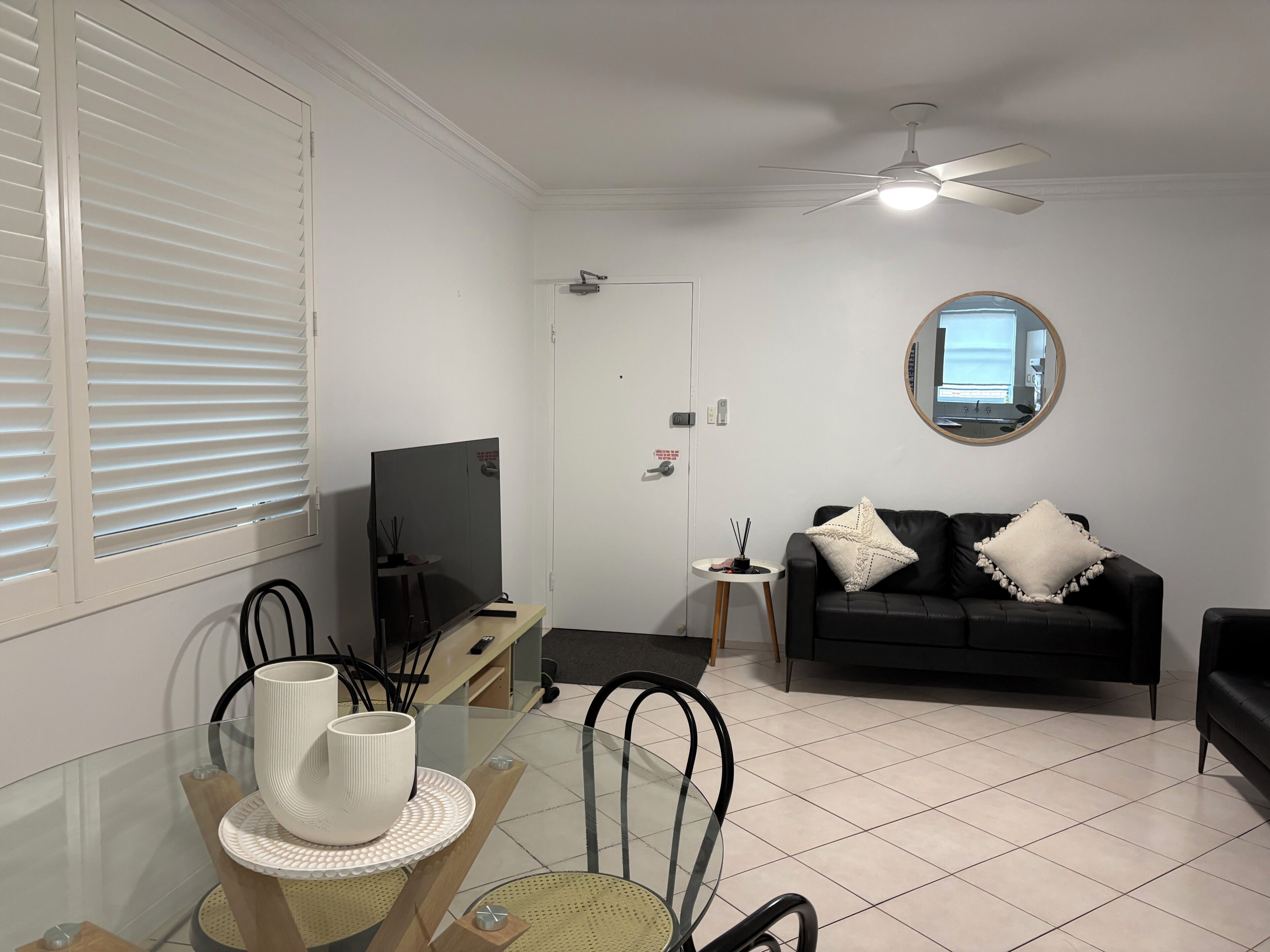 A living room with a TV, a black sofa, a round mirror, and a dining area with chairs and a table.