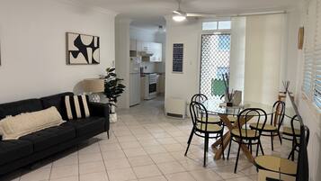 A living room with a black sofa, a dining table with chairs, a ceiling fan, and a kitchen in the background.