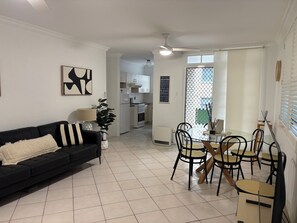 Un salon avec un canapé noir, une table à manger avec des chaises, un ventilateur de plafond et, au fond, une cuisine.