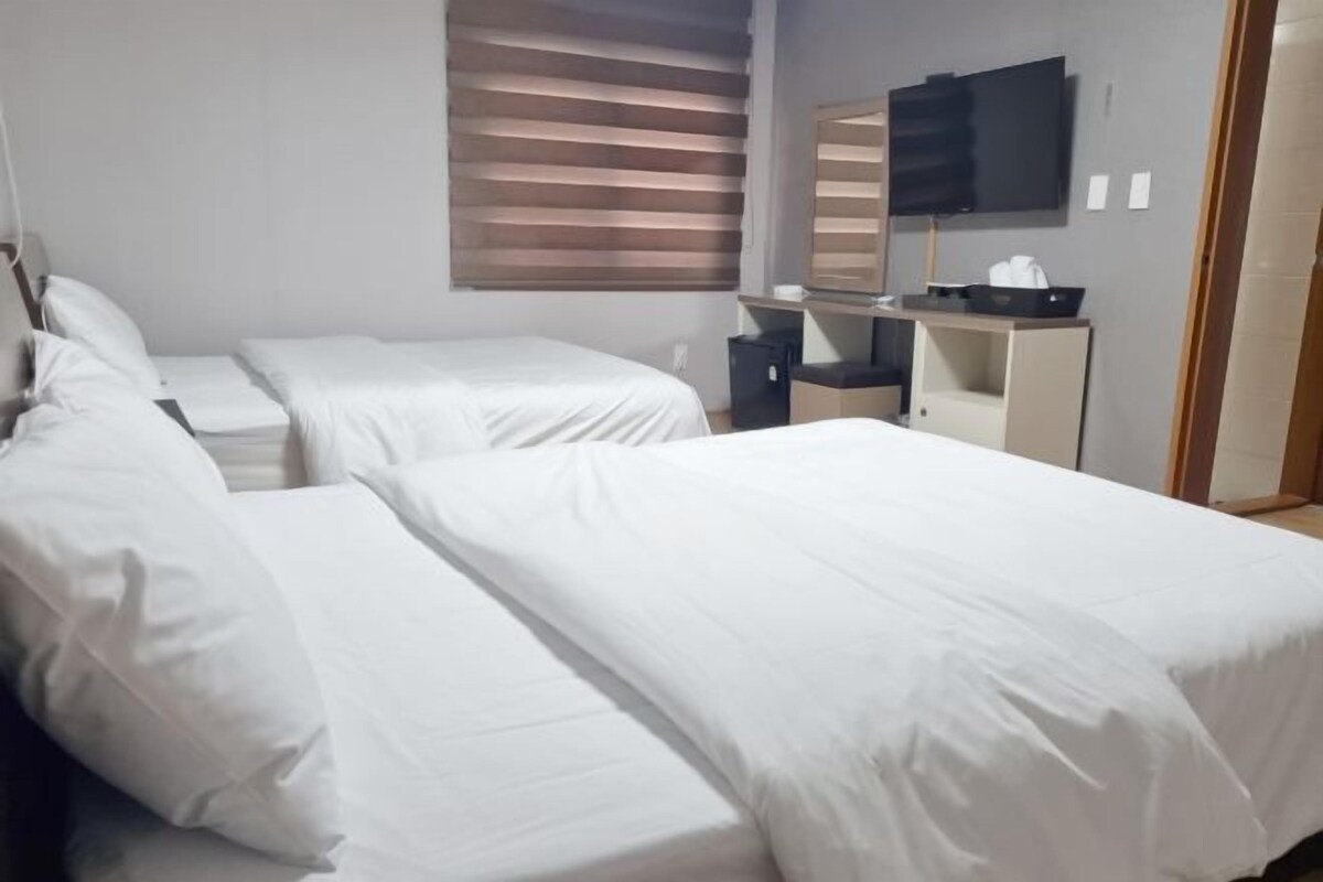 A hotel room with two single beds, a desk, a chair, a television, and a window with blinds.