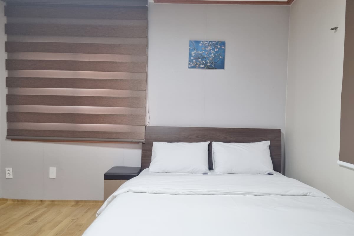 A bedroom with a bed, a nightstand, a window with blinds, and a small painting on the wall.