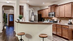 A modern kitchen with wooden cabinets, a stainless steel refrigerator, a microwave, and a coffee maker. There are two stools and a potted plant on the counter. - Lxry 4 BR, Heated Pool/Spa, Large Game Room, near Seaworld/Six Flags (St1) (San Antonio)
