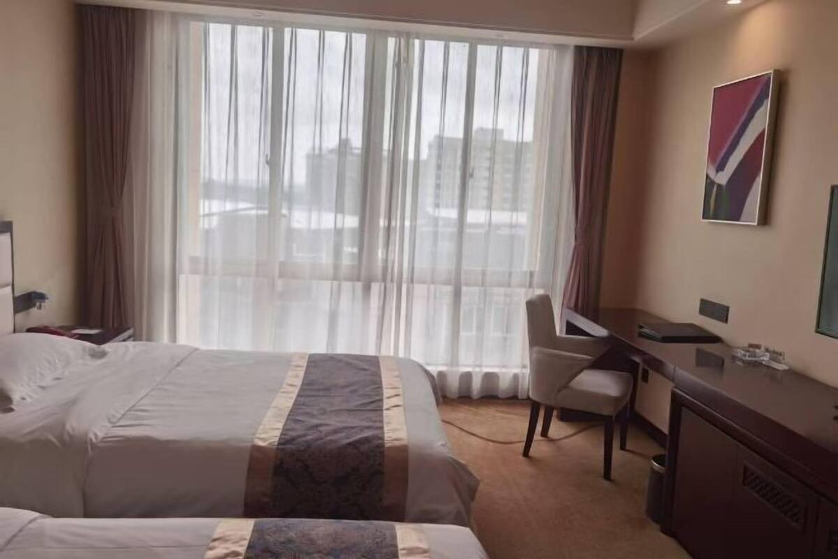 Habitación de hotel con cama, escritorio, silla y un amplio ventanal con cortinas.