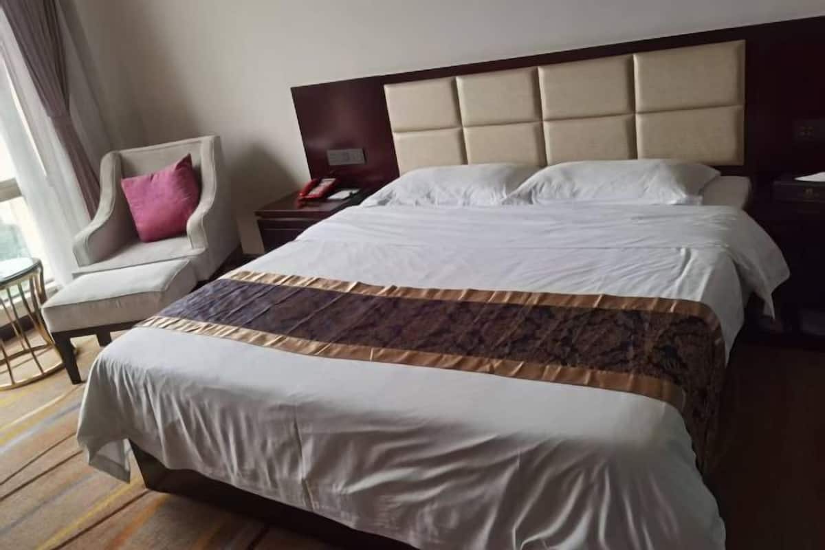 Habitación de hotel con una cama grande, una silla con un cojín rosa, una mesita de noche y una ventana con cortinas.