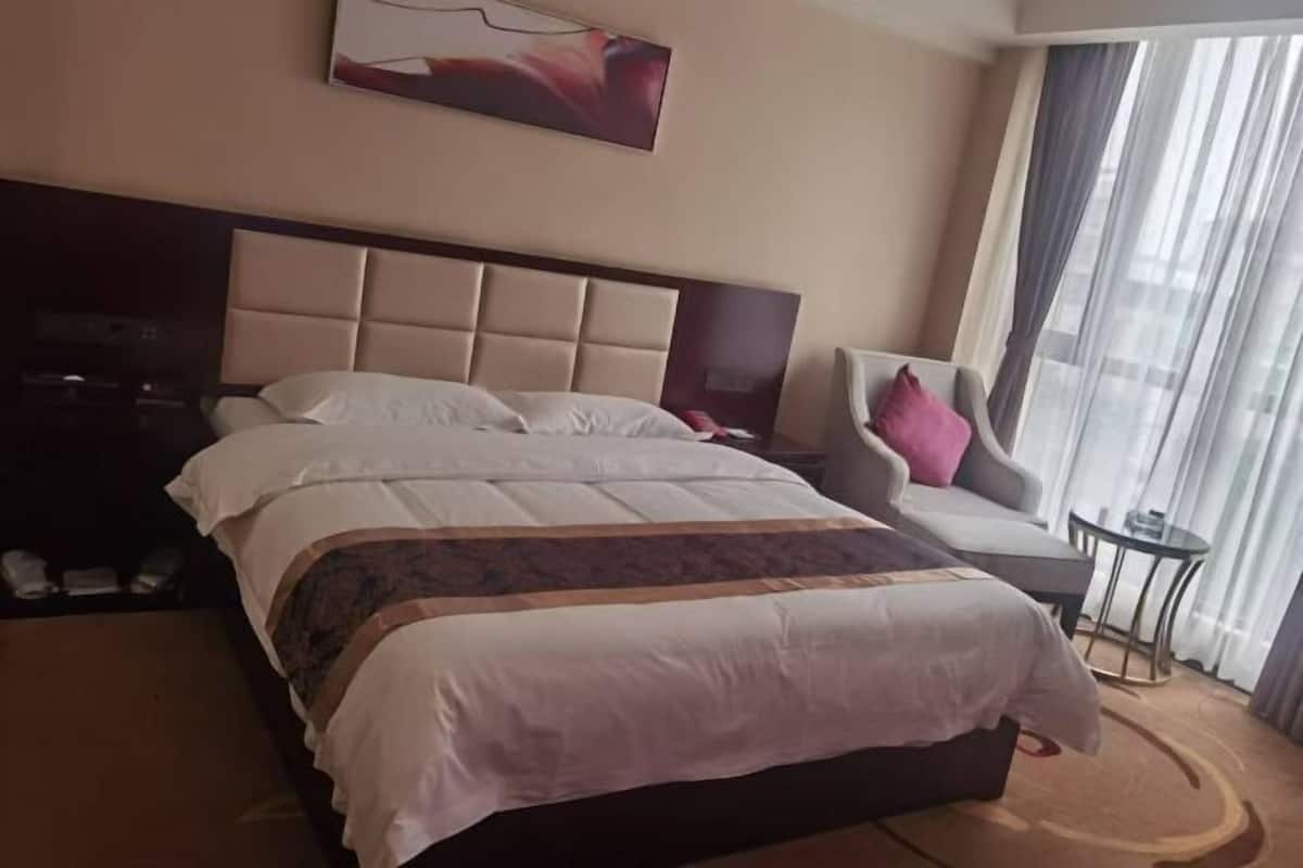 Habitación de hotel con una cama grande, cabecero, una silla, una mesita y un ventanal con cortinas.