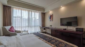 A hotel room with a large bed, a desk with a flat-screen TV, a chair, and a small table.