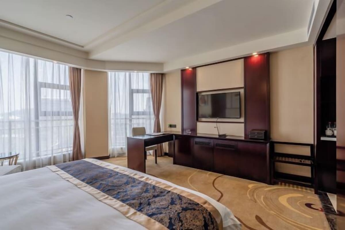 Habitación de hotel con una cama grande, un escritorio, televisión y vistas a la ciudad a través de amplias ventanas.