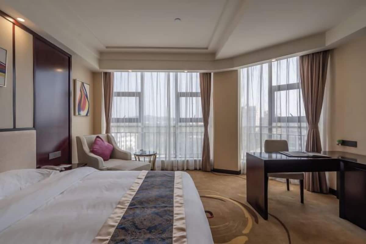 Habitación de hotel con una cama grande, un escritorio, una silla, una mesita y vistas a la ciudad a través de amplios ventanales.