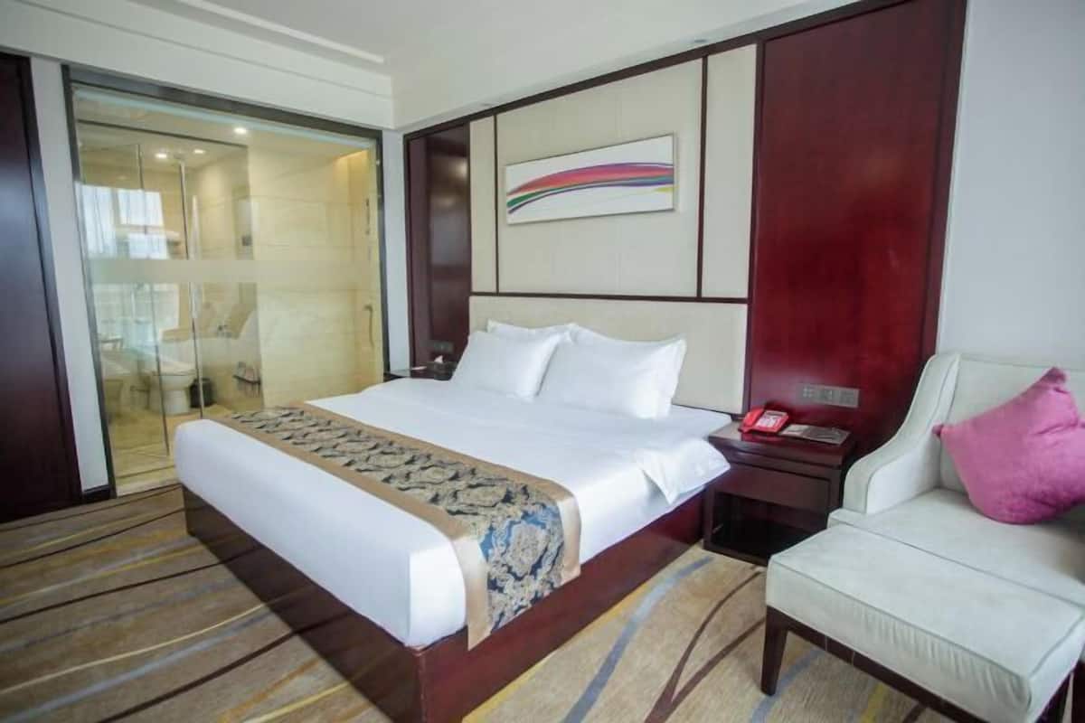 Habitación de hotel con una cama grande, una mesita de noche con teléfono, una silla y una puerta de vidrio que da al baño.