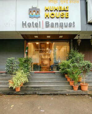 Indgangen til Mumbai House Hotel med trapper op til indgangen, plantekasser på hver side og et skilt over indgangen.