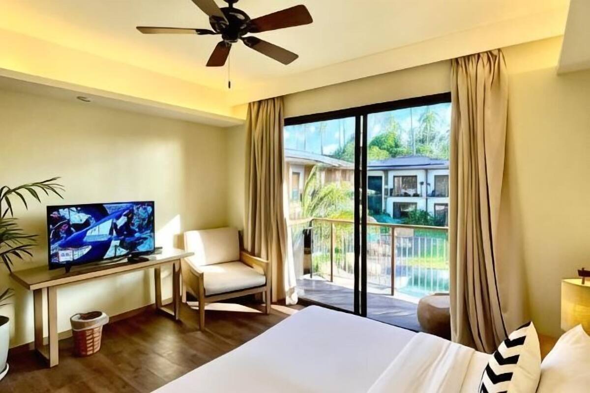 Ein Hotelzimmer mit Bett, Fernseher, Schreibtisch, Stuhl, Deckenventilator und einem Balkon mit Blick ins Grüne.