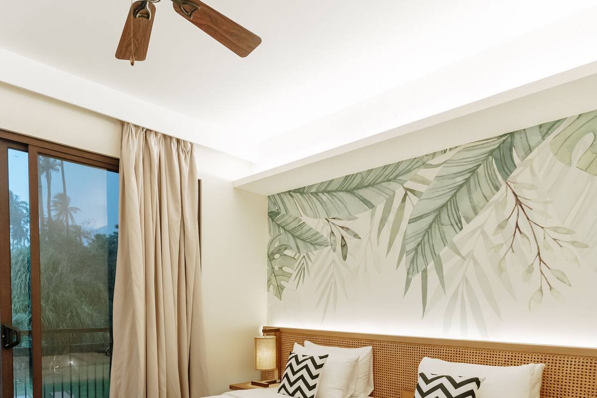 Ein Hotelzimmer mit zwei Betten, einem Deckenventilator, einem großen Fenster mit Vorhängen und einem Wandgemälde mit tropischen Blättern.