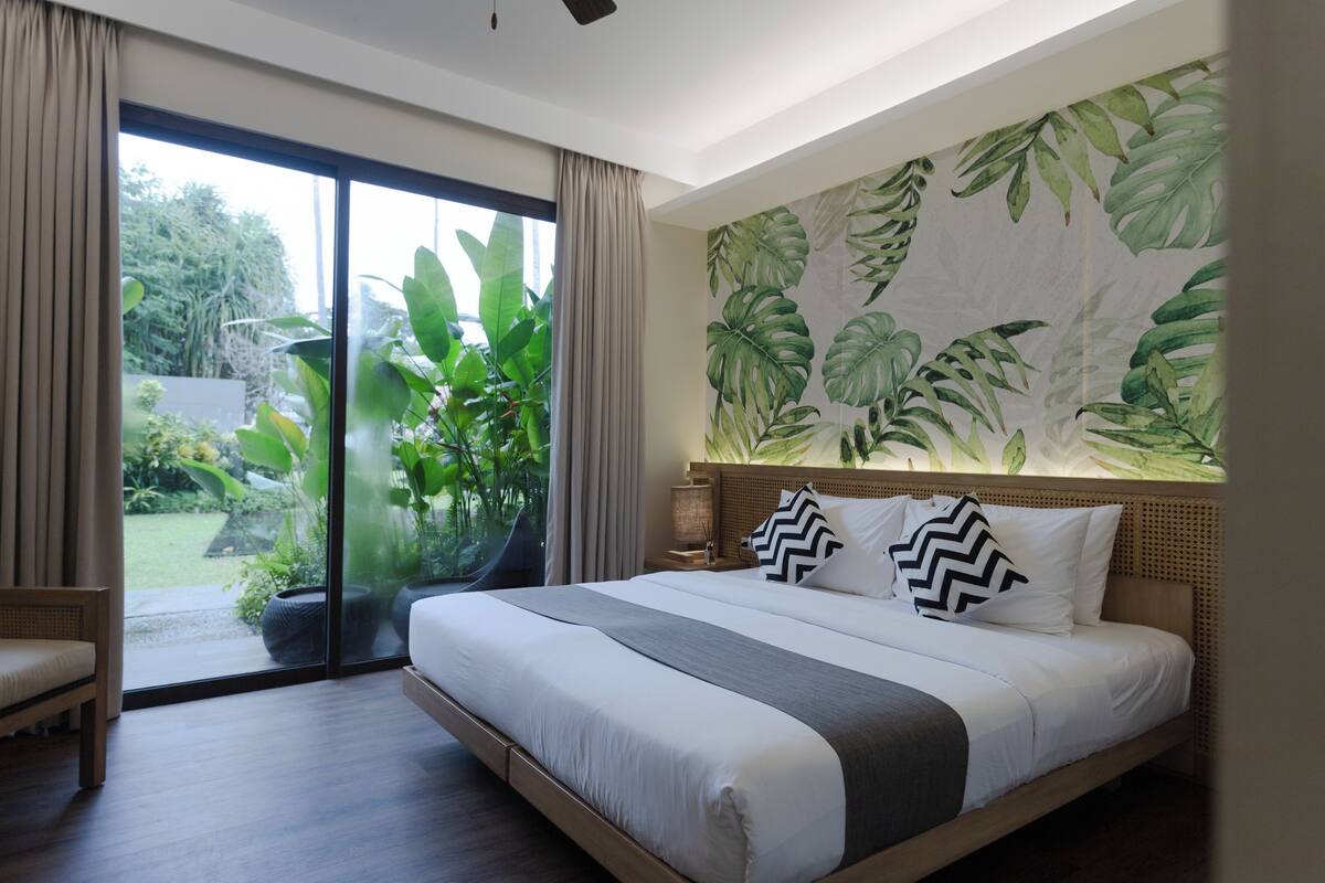 Ein Schlafzimmer mit einem großen Bett, Blick ins Grüne durch eine Schiebetür und einer Wand mit tropischem Blattmuster.