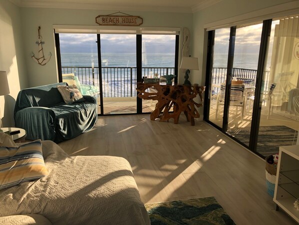A room with a large sliding glass door leading to a balcony with a view of the ocean. There is a sofa and a small table with decorative items.