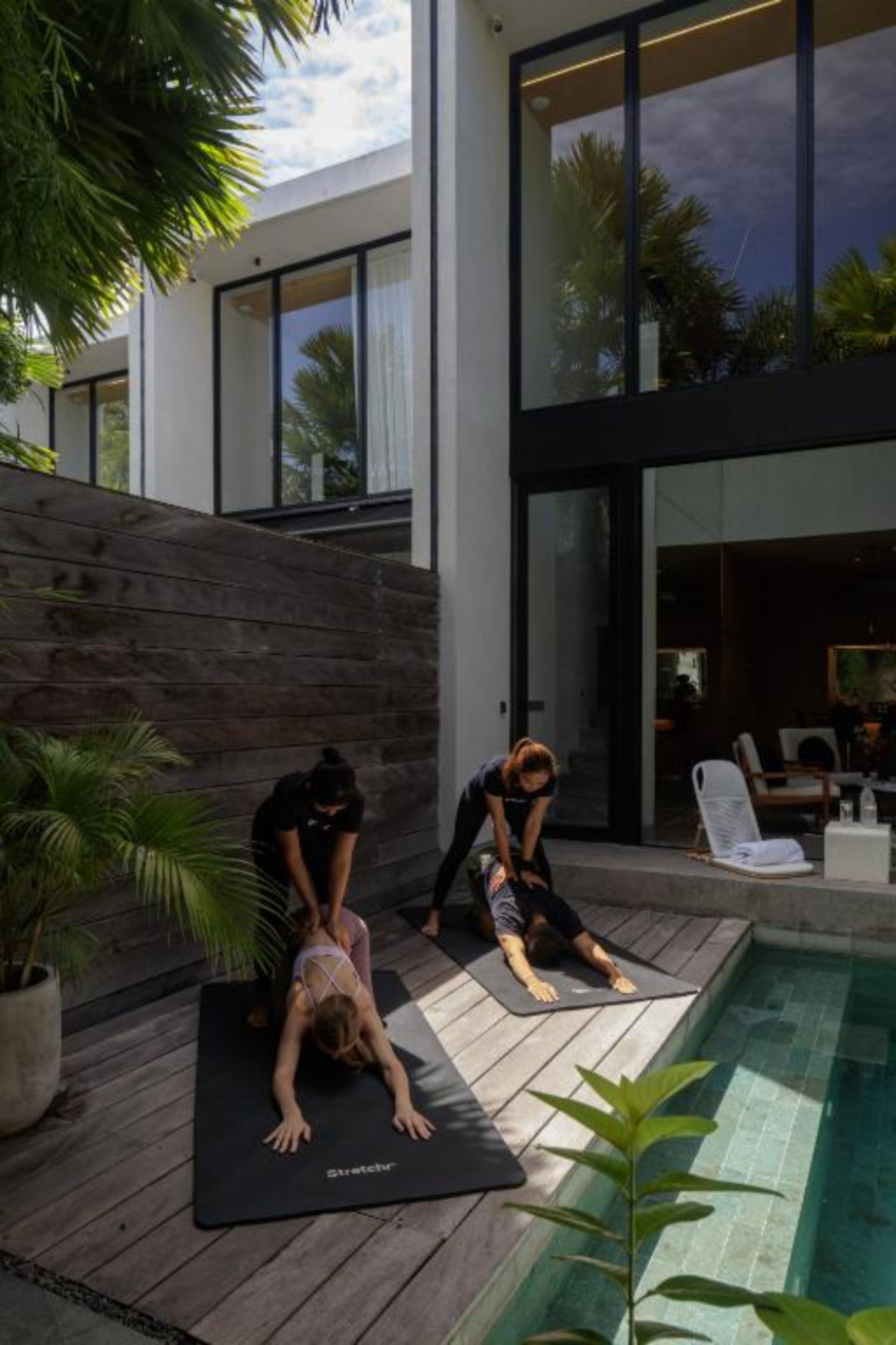 Un'area yoga all'aperto moderna con un pavimento in legno, una piscina e un edificio in vetro sullo sfondo.