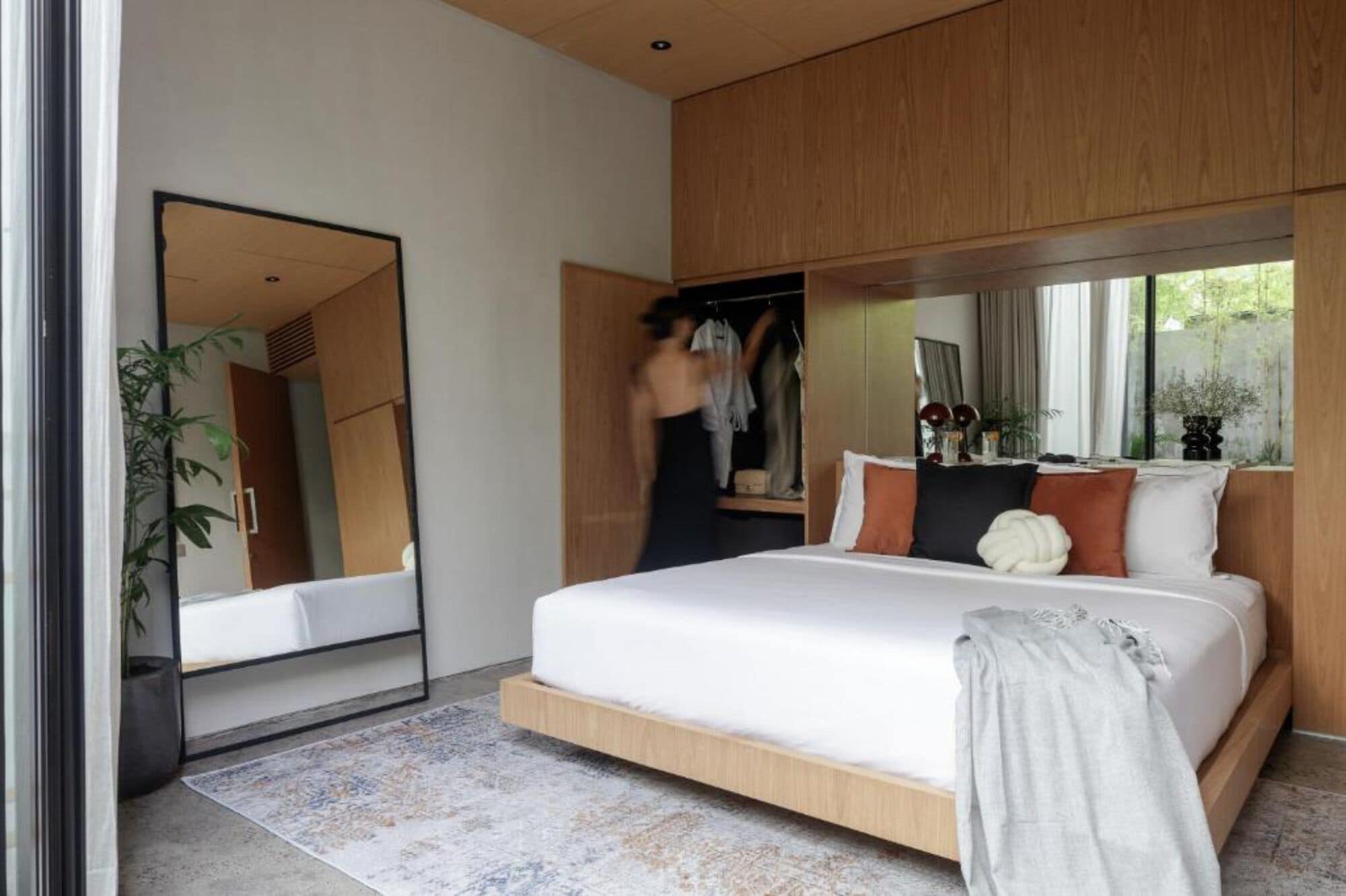 A modern bedroom with a large bed, a wardrobe, a mirror, and a view of greenery outside.
