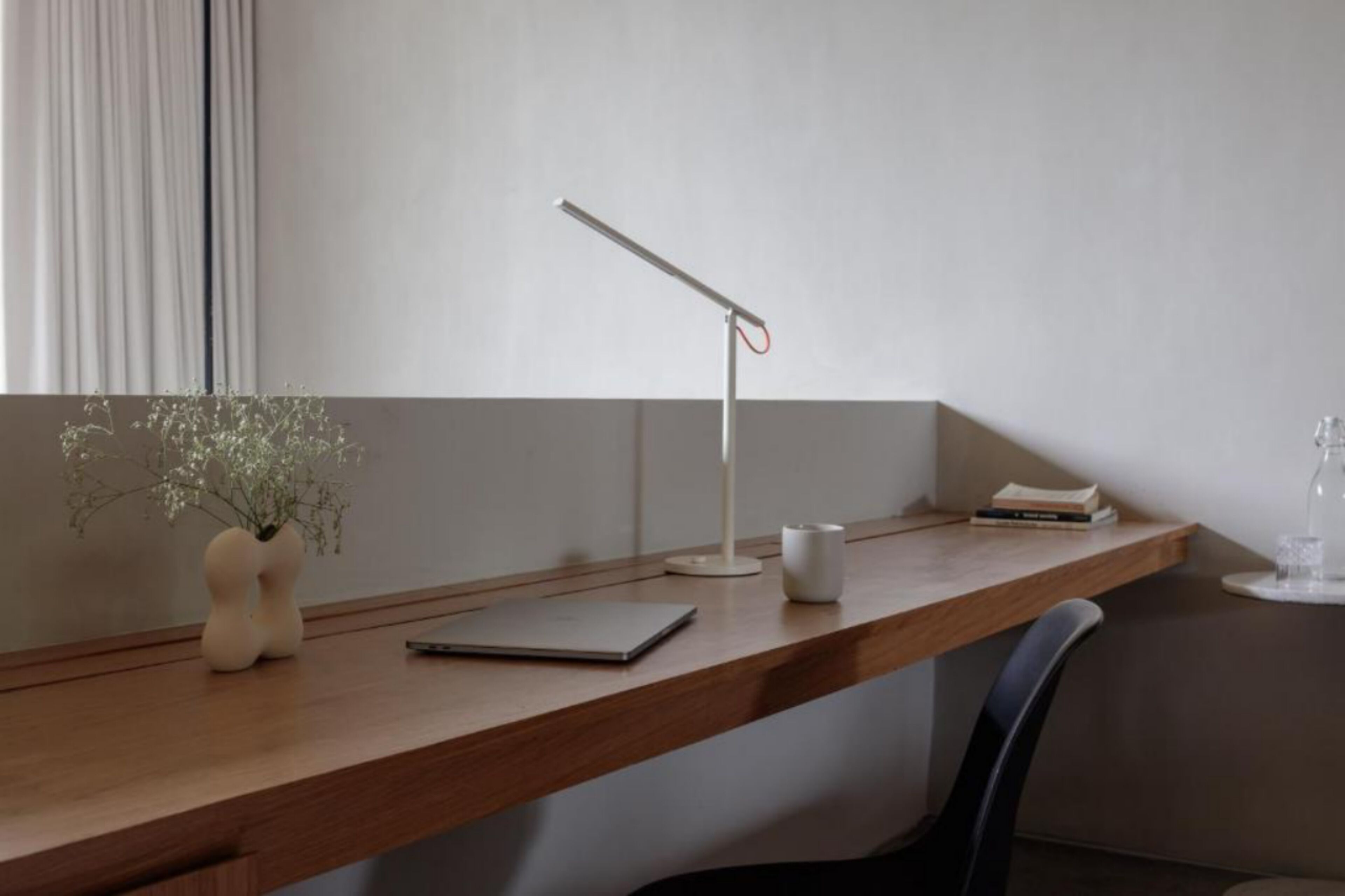 A wooden desk with a white desk lamp, a vase with flowers, a notebook, and a glass of water.