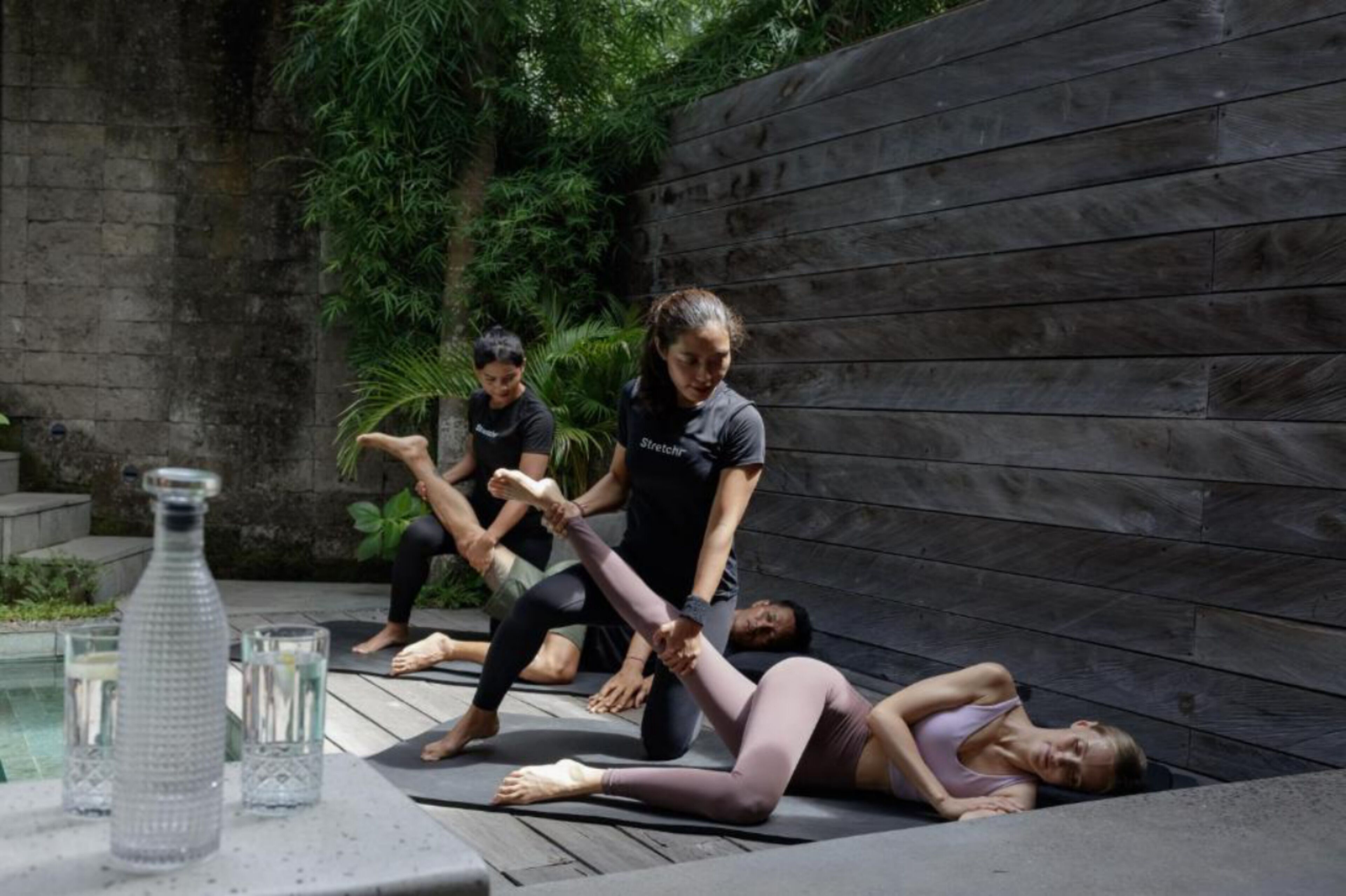A group of people performing a stretching exercise outdoors near a wooden wall and a pool.