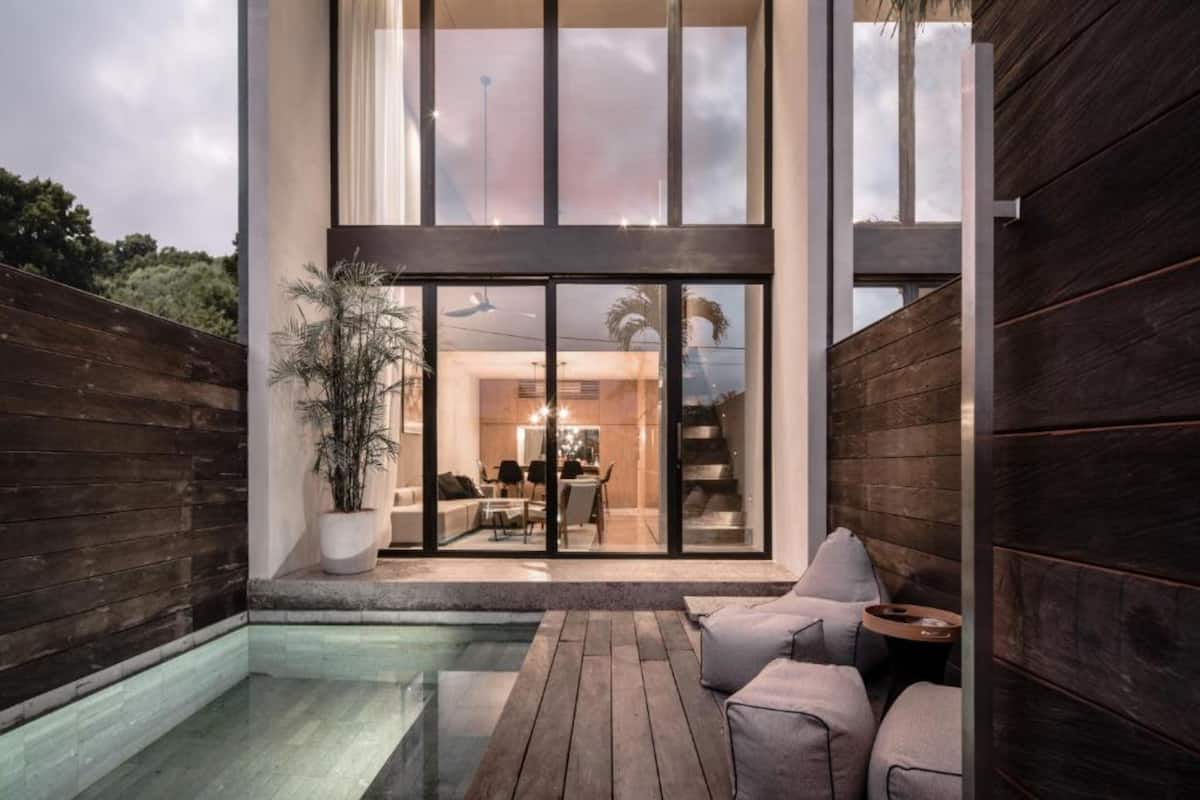 Een modern huis met een grote glazen deur die uitkomt op een eethoek, een zwembad en een houten terras.
