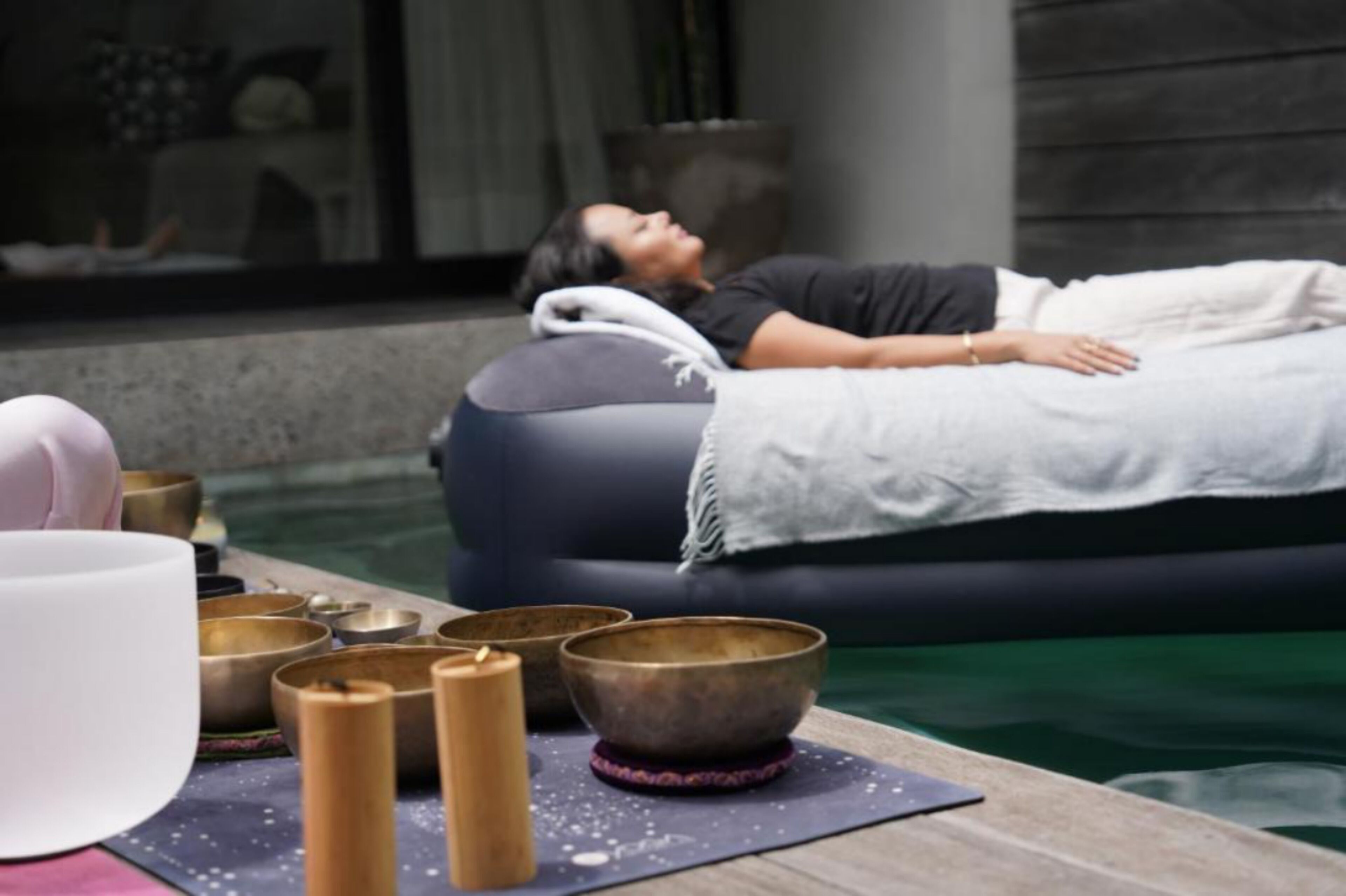 Iemand ontspant op een drijvend bed bij een zwembad, met meditatiekommen en kaarsen.