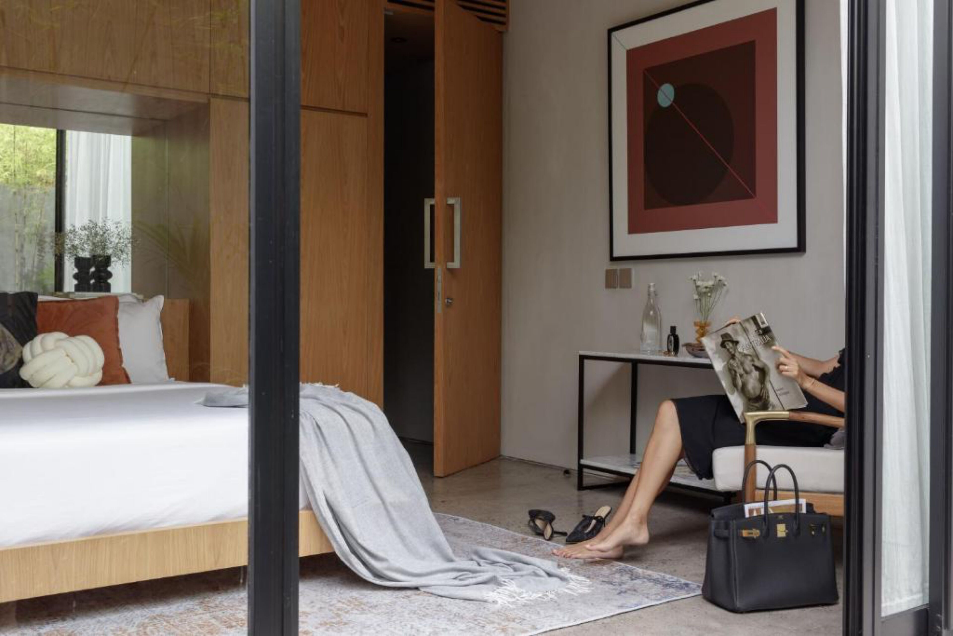 Une chambre moderne avec un lit, un fauteuil de lecture, une petite table surmontée d’un vase et, au mur, un tableau abstrait encadré.