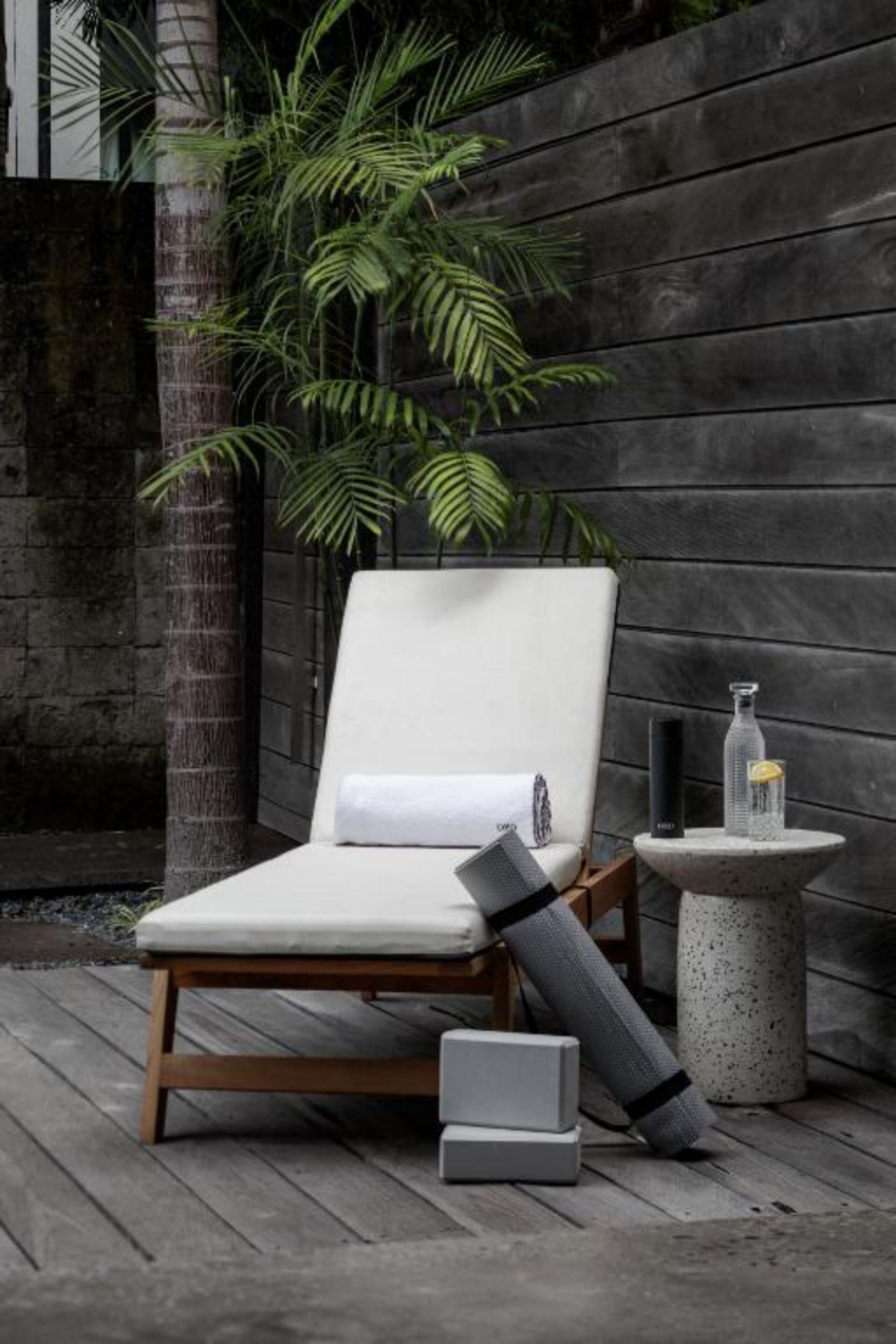 A lounge chair with a cushion, a small table with a bottle, and a rolled-up mat on a wooden deck.