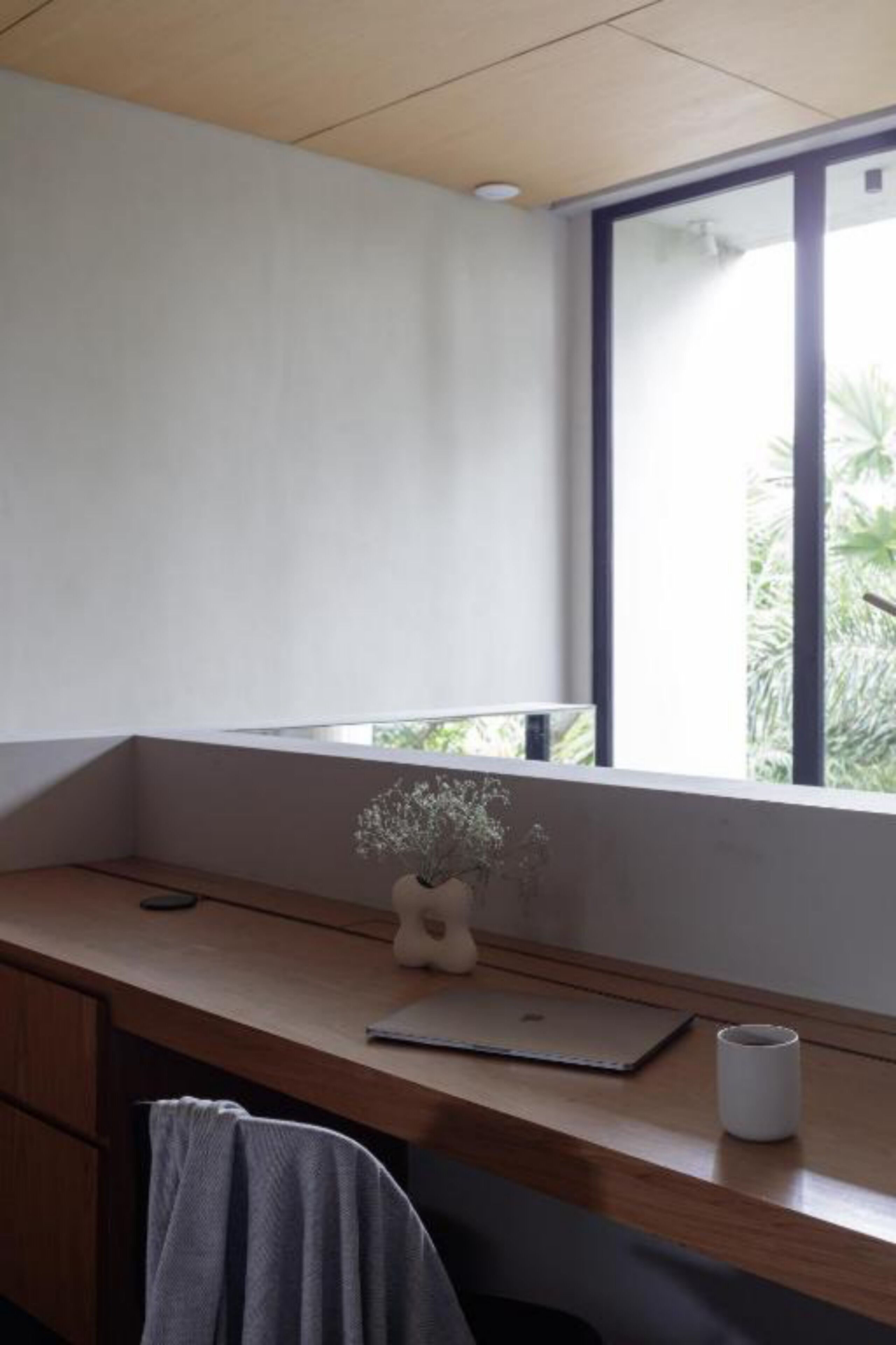 Quarto com escrivaninha de madeira, laptop, um vaso com flores e janela com vista para o verde.