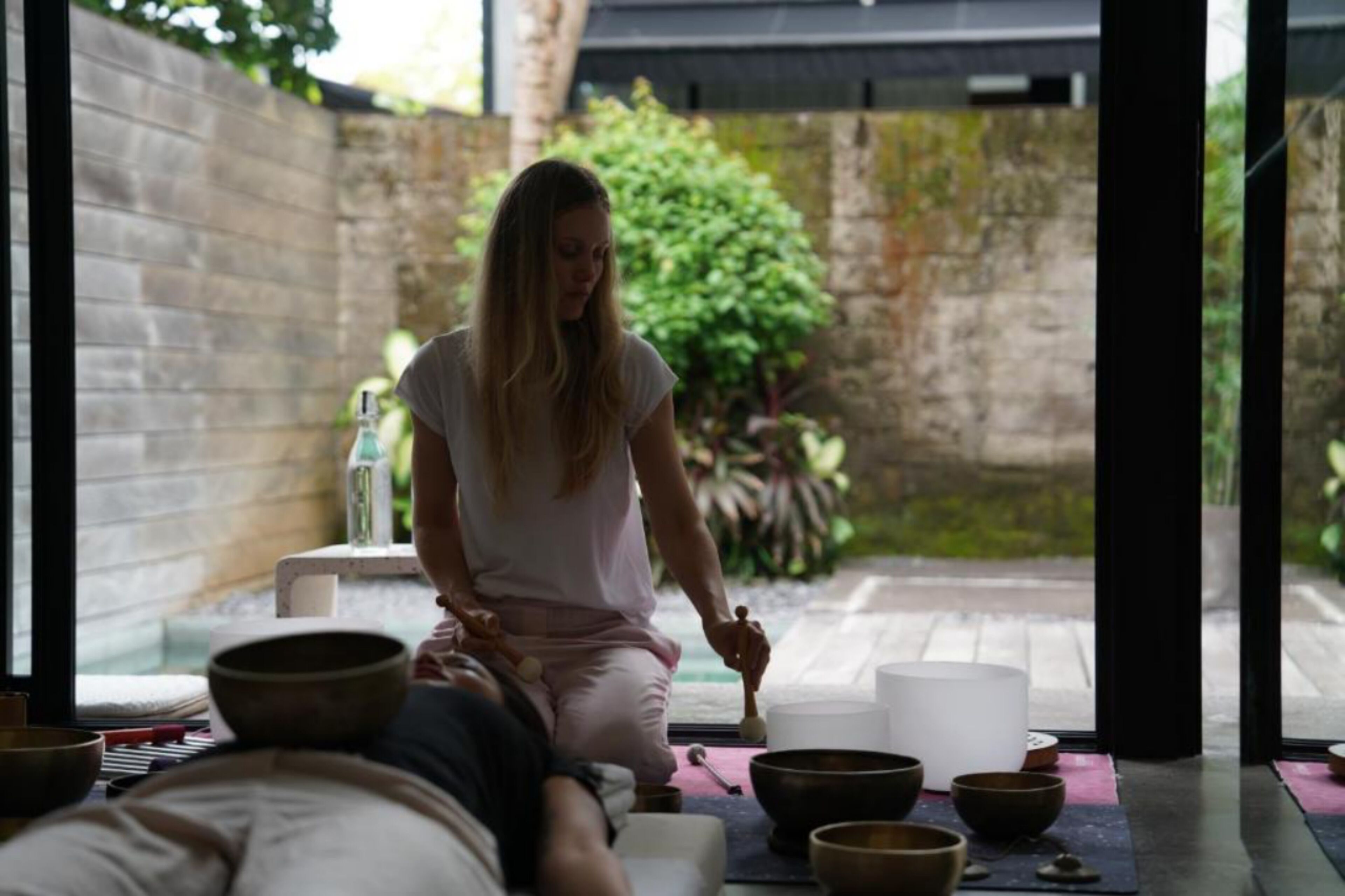 Una persona meditando en una habitación con vistas a un jardín, rodeada de cuencos y una botella de agua.