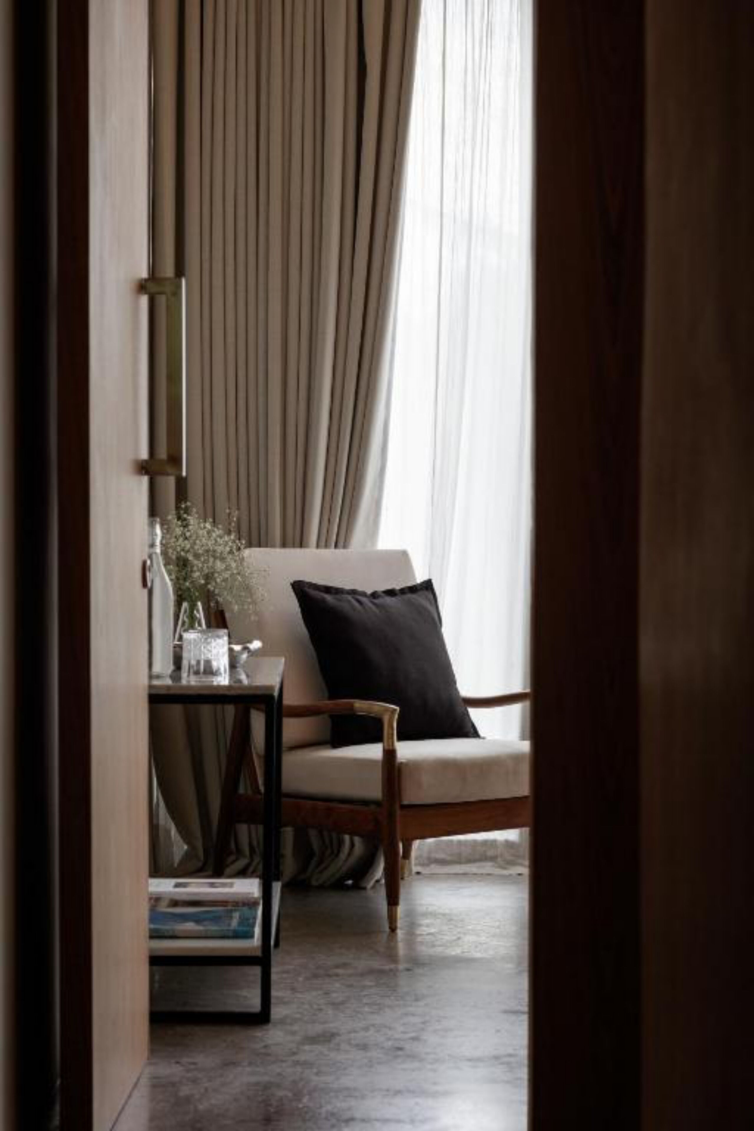 Een gezellige kamer met een houten stoel, een tafeltje met een vaas en boeken, en uitzicht op een raam met gordijnen.