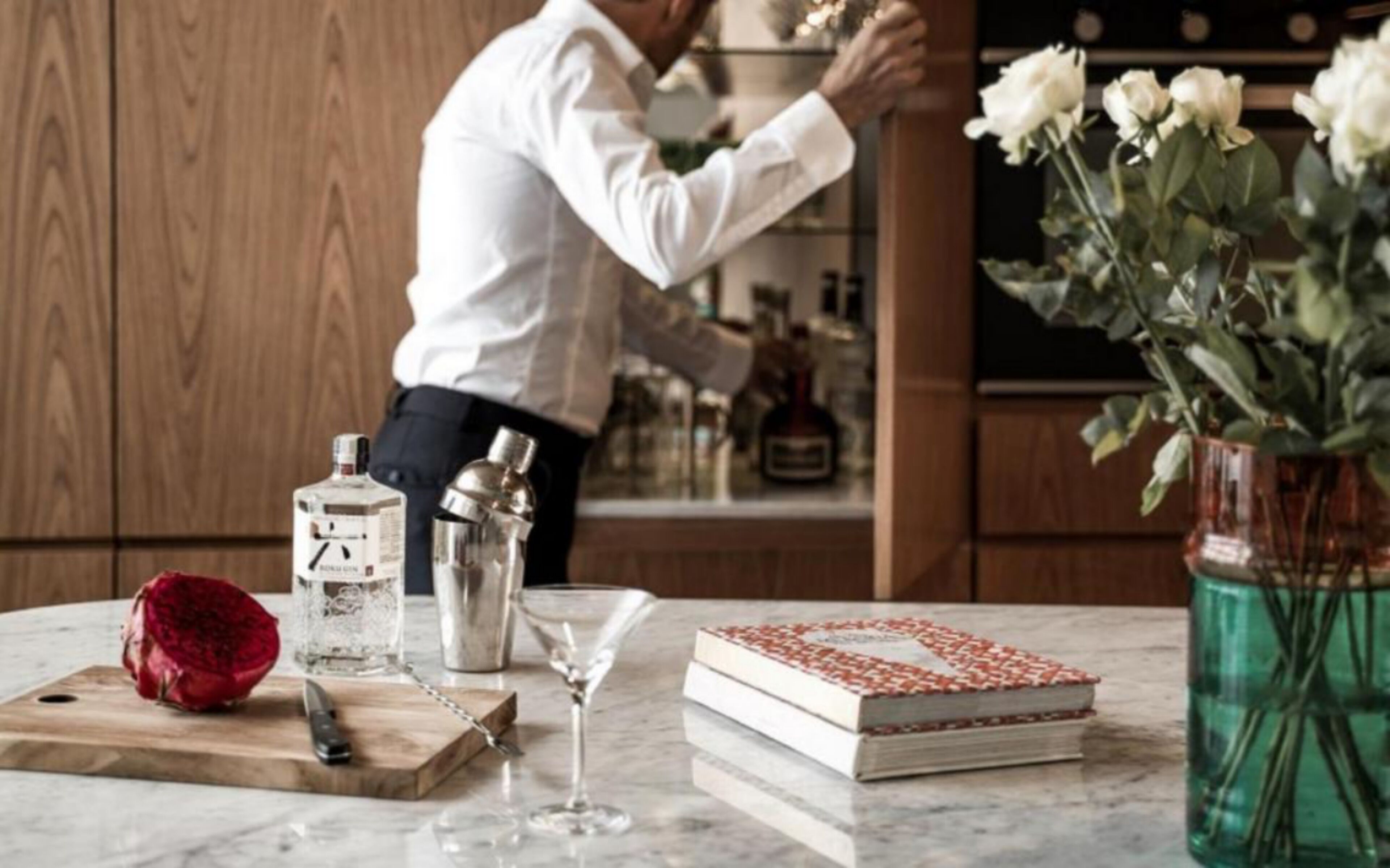 Un comptoir de cuisine avec une planche à découper, un livre, un verre à martini, une bouteille de spiritueux et un vase rempli de fleurs.