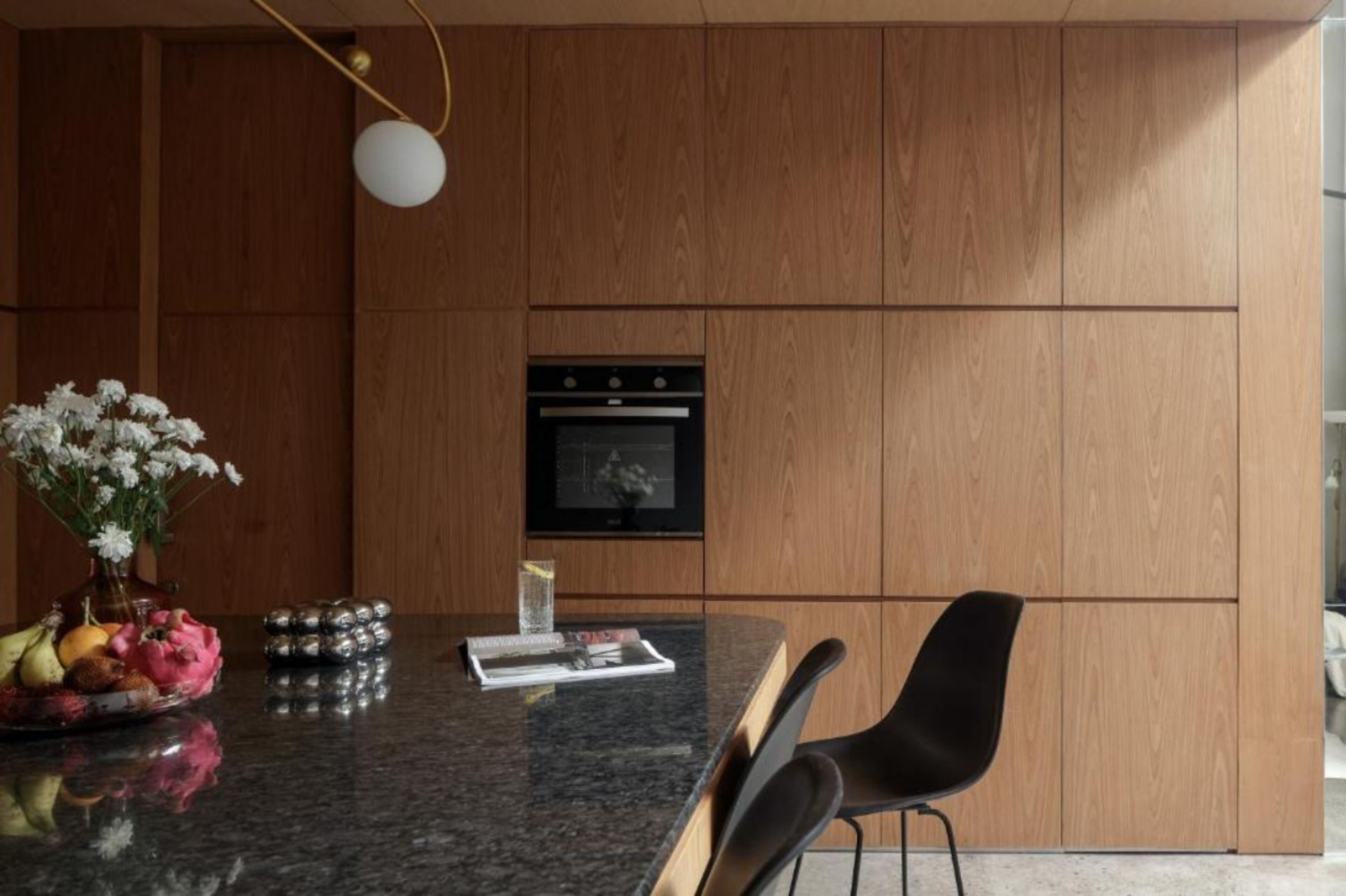 A modern kitchen with wooden cabinets, a built-in oven, a granite countertop, and a vase with flowers.