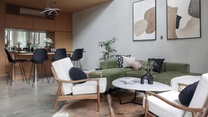 Un salon moderne avec un canapé vert, des fauteuils blancs et une table basse. Des tableaux abstraits ornent le mur, et on aperçoit une cuisine au fond.