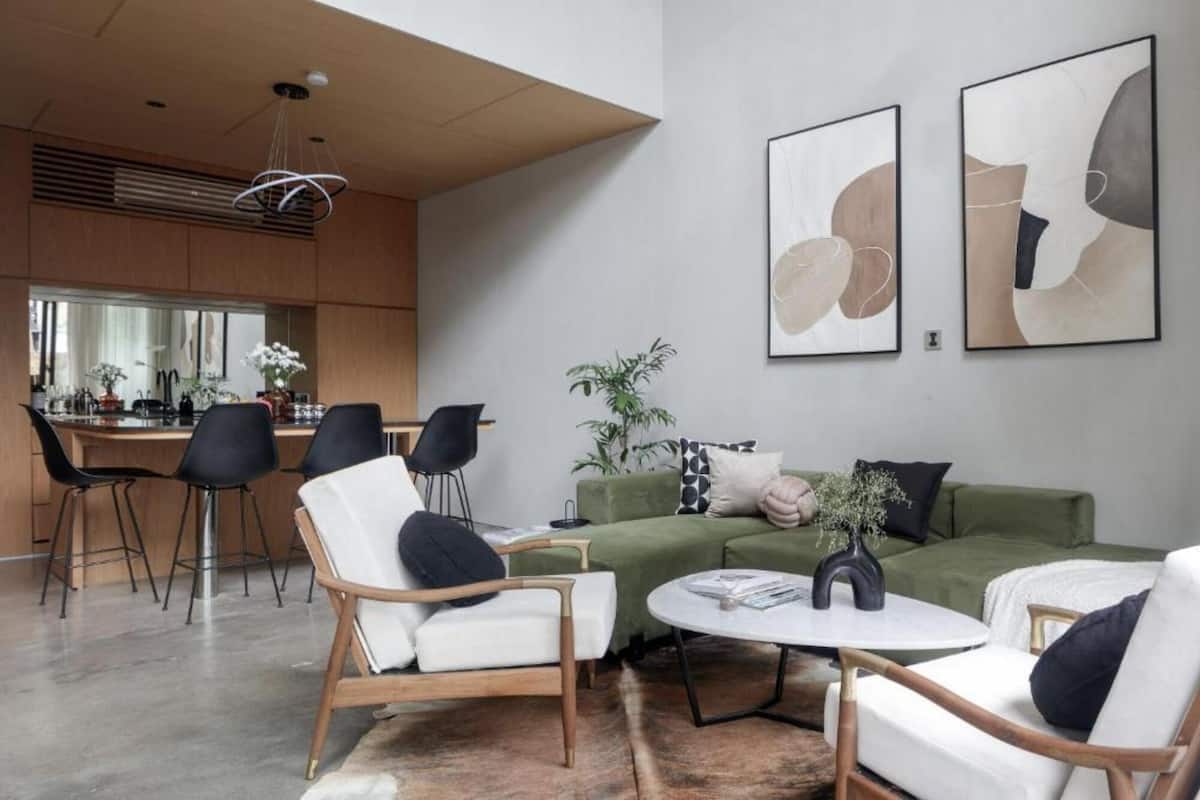 Een moderne woonkamer met een groene bank, witte fauteuils en een salontafel. Aan de muur hangen abstracte schilderijen en er is een keuken op de achtergrond.