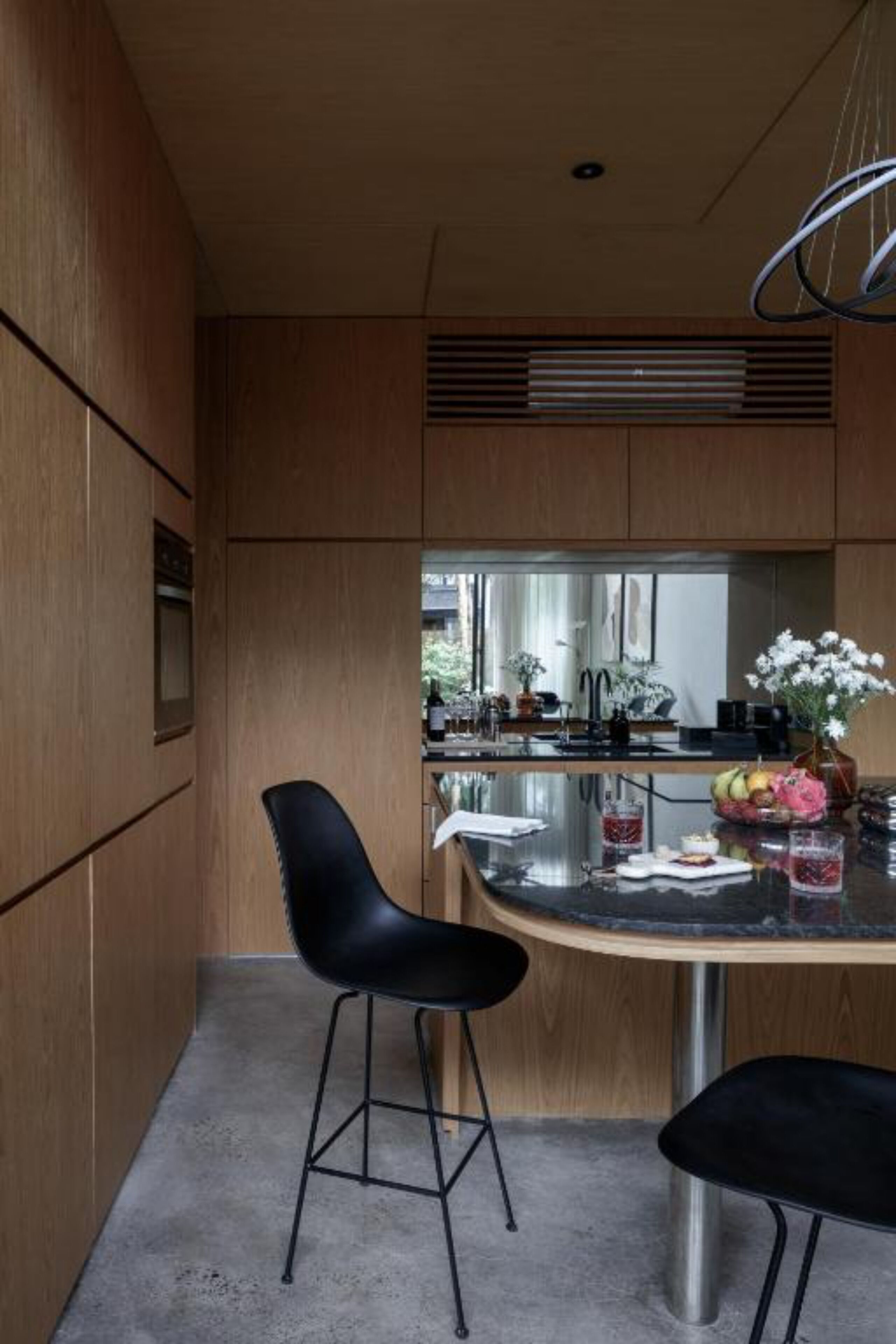 A modern kitchen with wooden cabinets, a black bar stool, a round table with a vase of flowers, and a hanging light fixture.