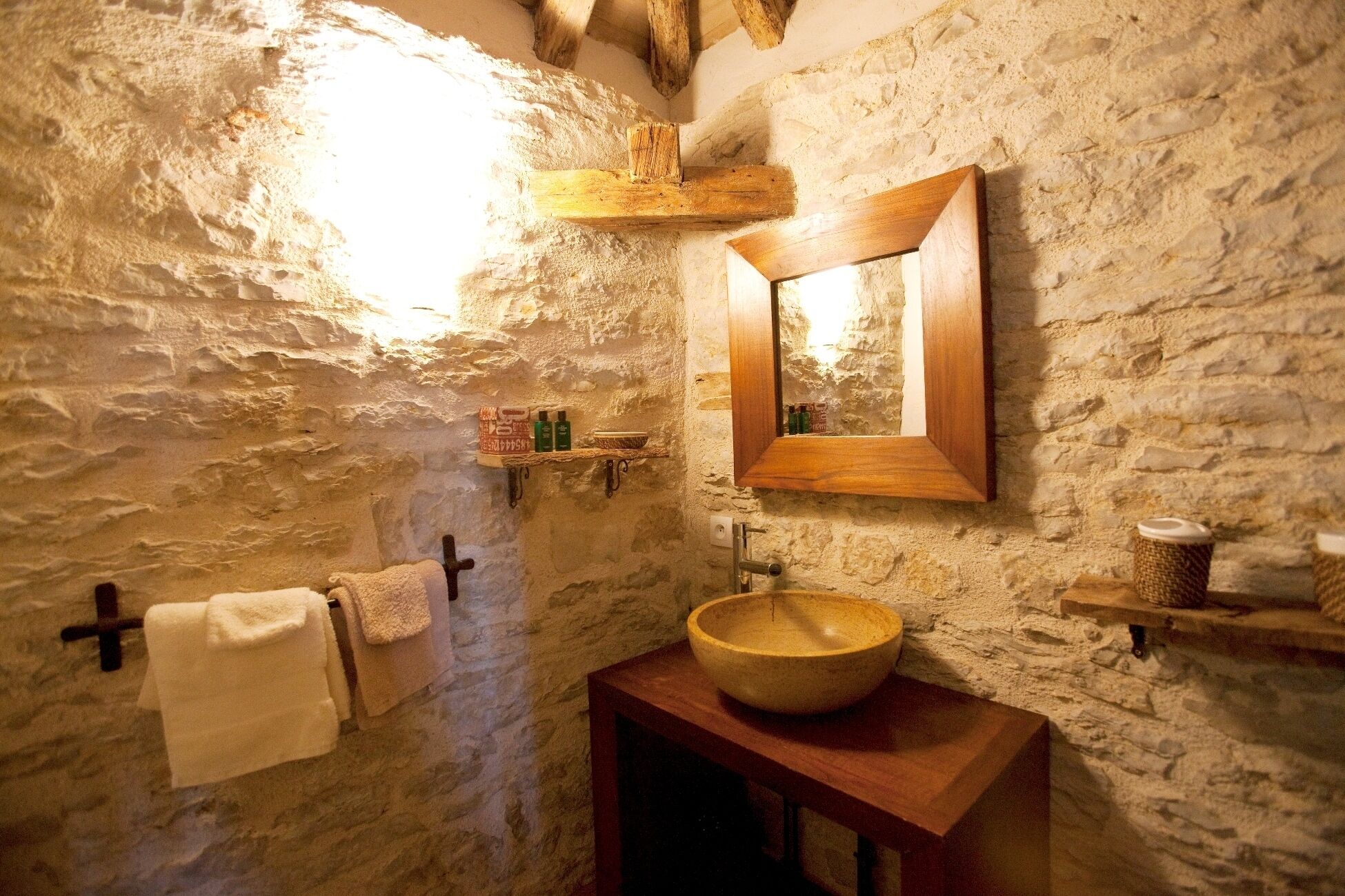 A rustic bathroom with a stone wall, wooden beams, a wooden basin, and a mirror.