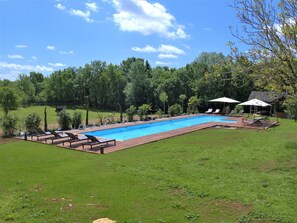 Une piscine rectangulaire avec une terrasse en bois, entourée de pelouse et d’arbres. On y trouve des chaises longues et des parasols sous un pavillon.