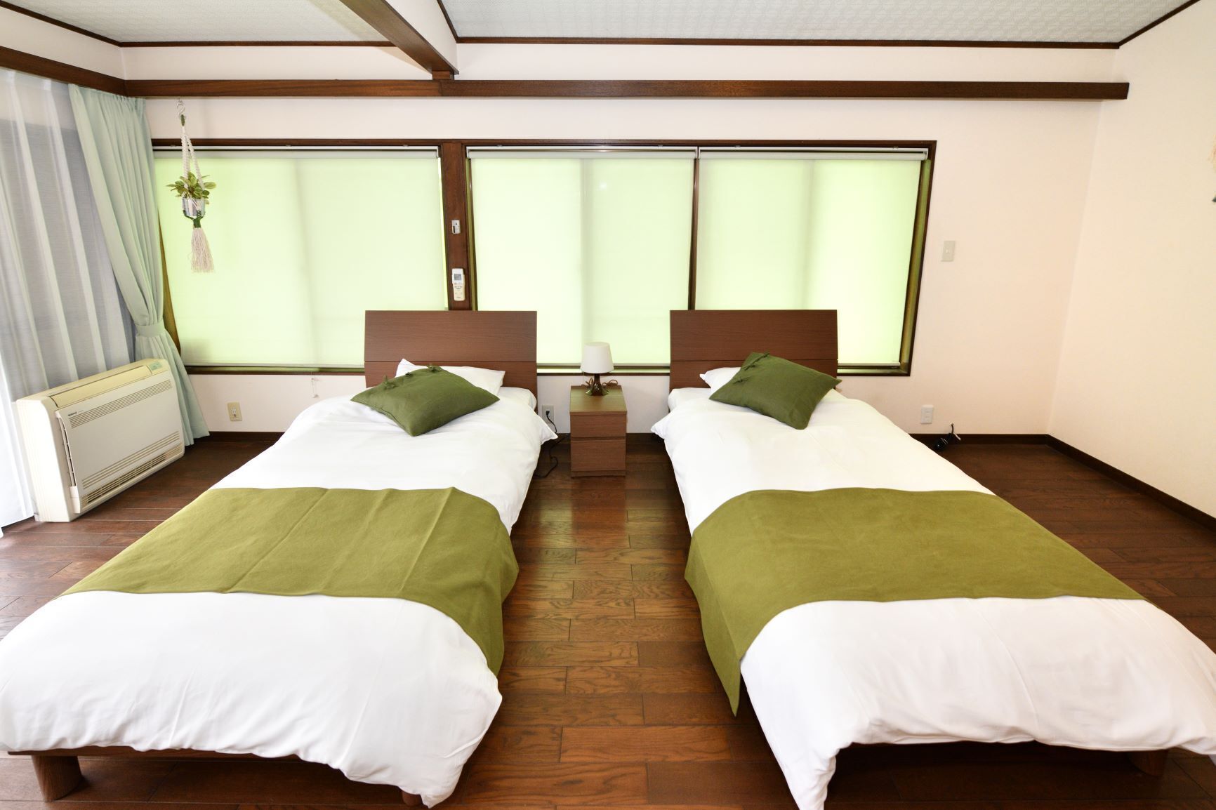 A twin room with two beds, each with a green pillow and white bedding, a bedside table, and a window with curtains.