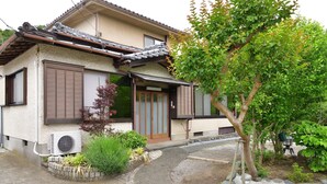 Uma casa tradicional japonesa com telhado de telhas, portas deslizantes e um pequeno jardim com plantas e uma árvore.