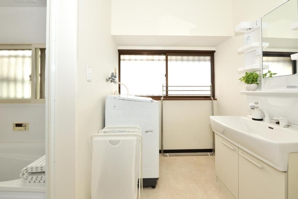 A compact hotel room with a washing machine, a sink, and a window with blinds.