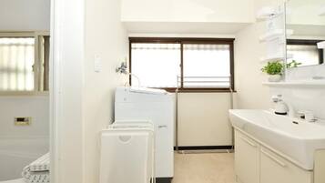 A compact hotel room with a washing machine, a sink, and a window with blinds.