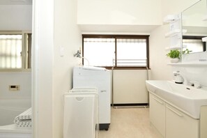 A compact hotel room with a washing machine, a sink, and a window with blinds. - wasabi (Kamakura)