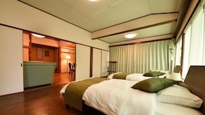 Quarto de hotel com cama grande, escrivaninha e uma cadeira. Há uma janela com cortinas e uma planta pendurada próximo à janela.