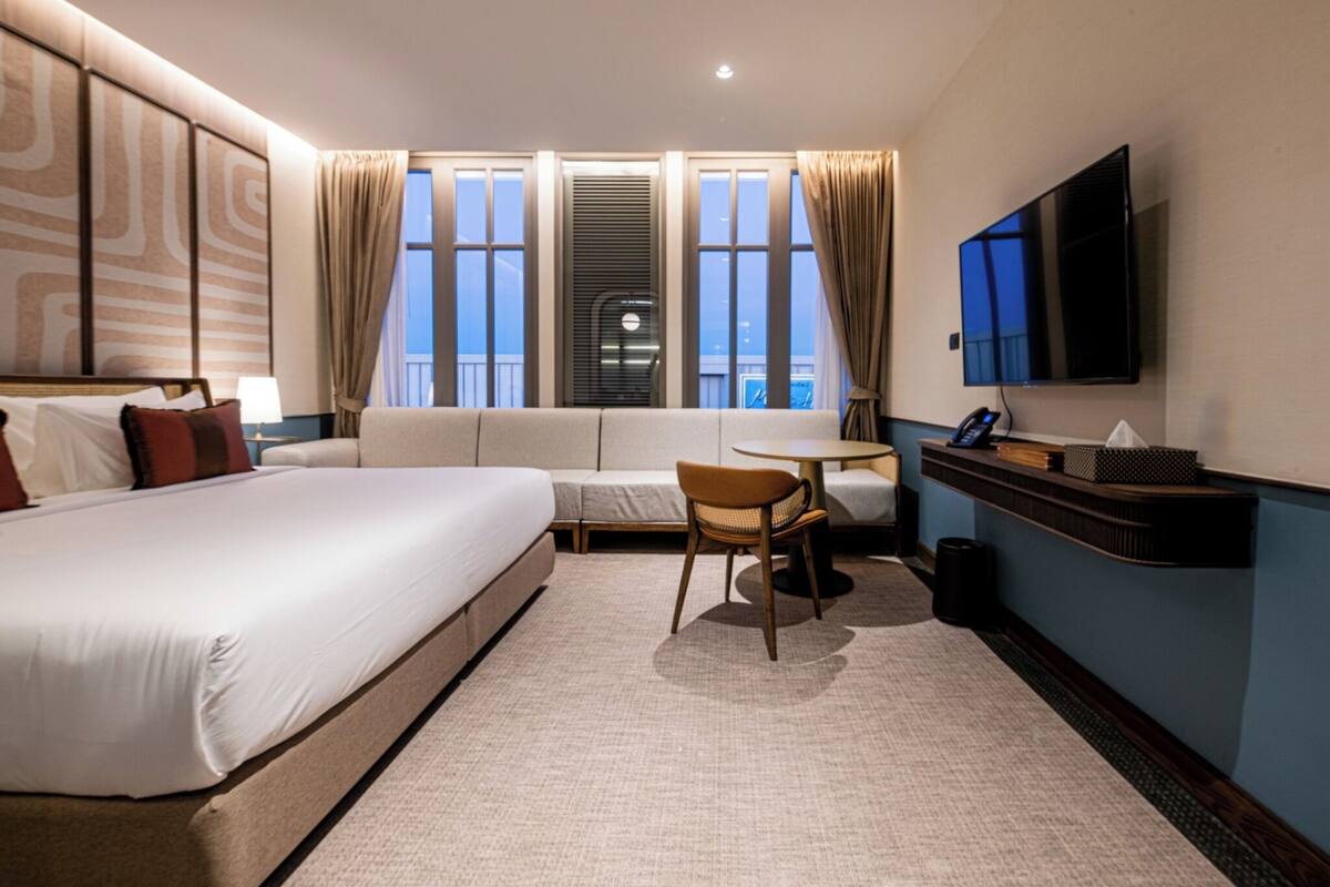 A hotel room with a large bed, a sofa, a small table, a chair, a TV, and a window with curtains.