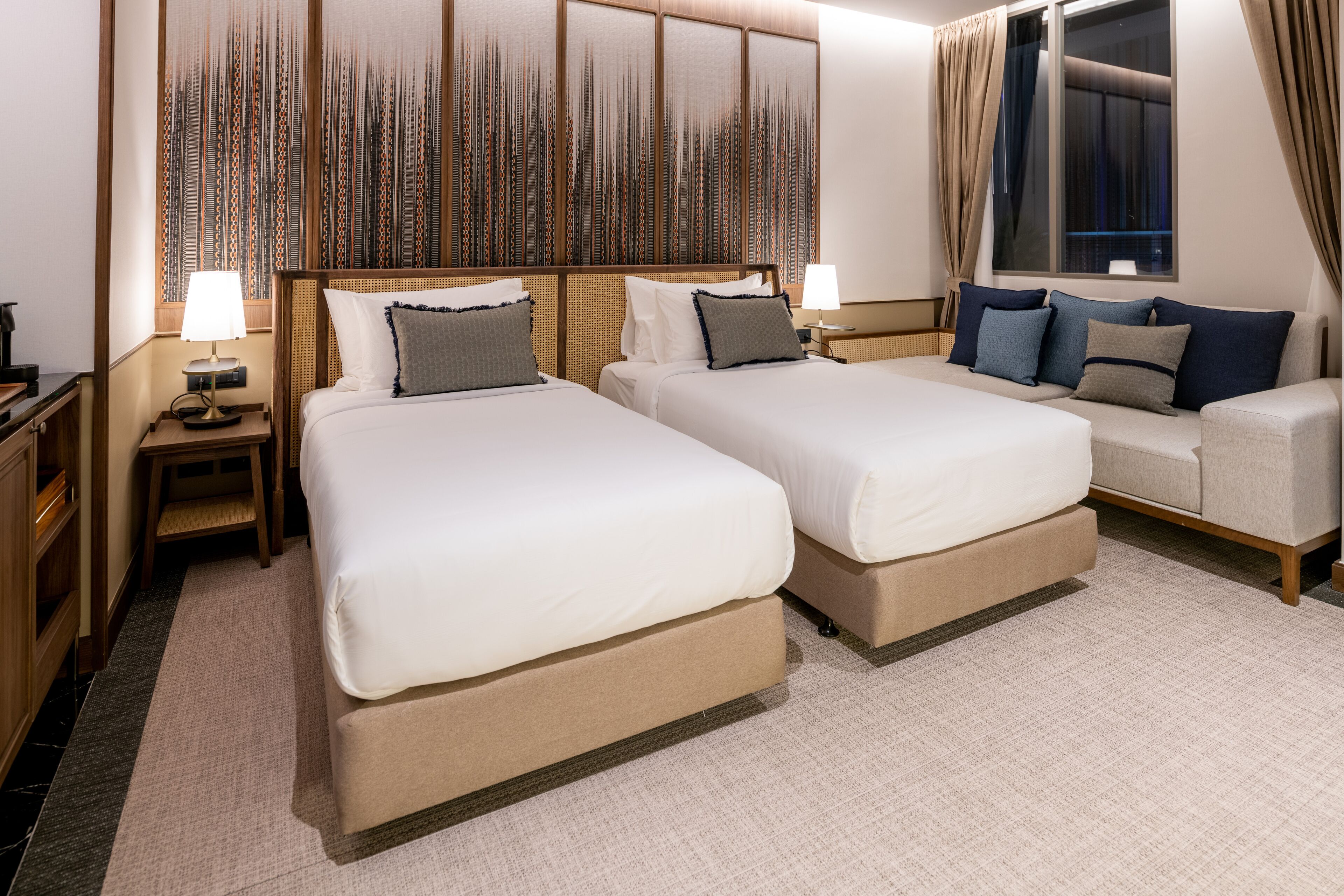 A hotel room with two beds, a sofa, a large window with curtains, and a decorative wall panel.
