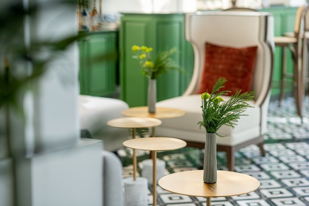 Una habitación con un aparador verde, un sillón blanco con cojín rojo y tres mesitas redondas con jarrones de follaje.