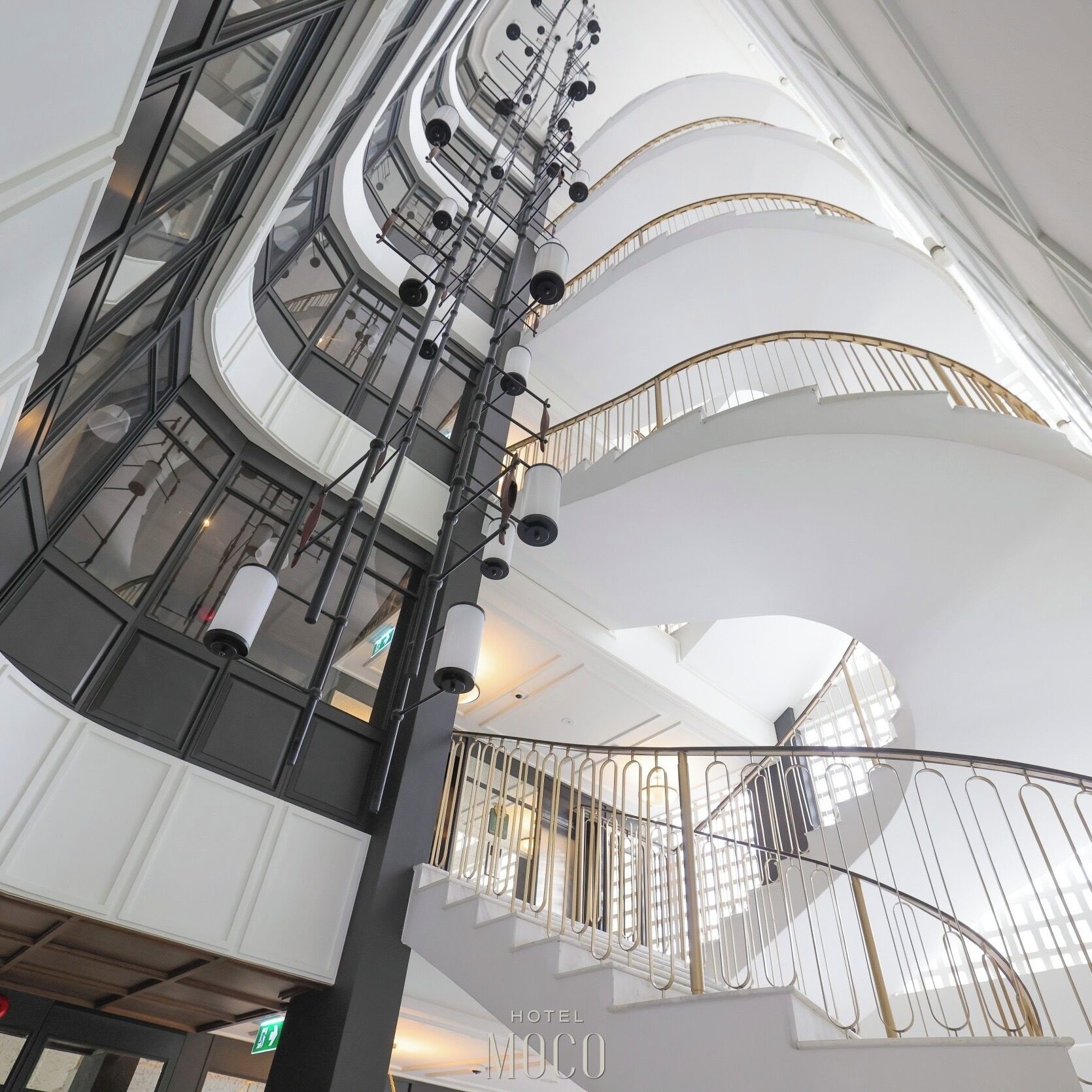 Interior moderno de un hotel con varios niveles de balcones, una escalera en espiral central y vistas del cielo a través de los pisos superiores.