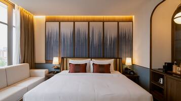 Una habitación de hotel con una cama grande, dos mesitas de noche con lámparas, un sofá y un ventanal con cortinas.