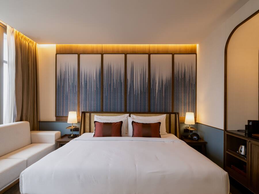 Una habitación de hotel con una cama grande, dos mesitas de noche con lámparas, un sofá y un ventanal con cortinas.