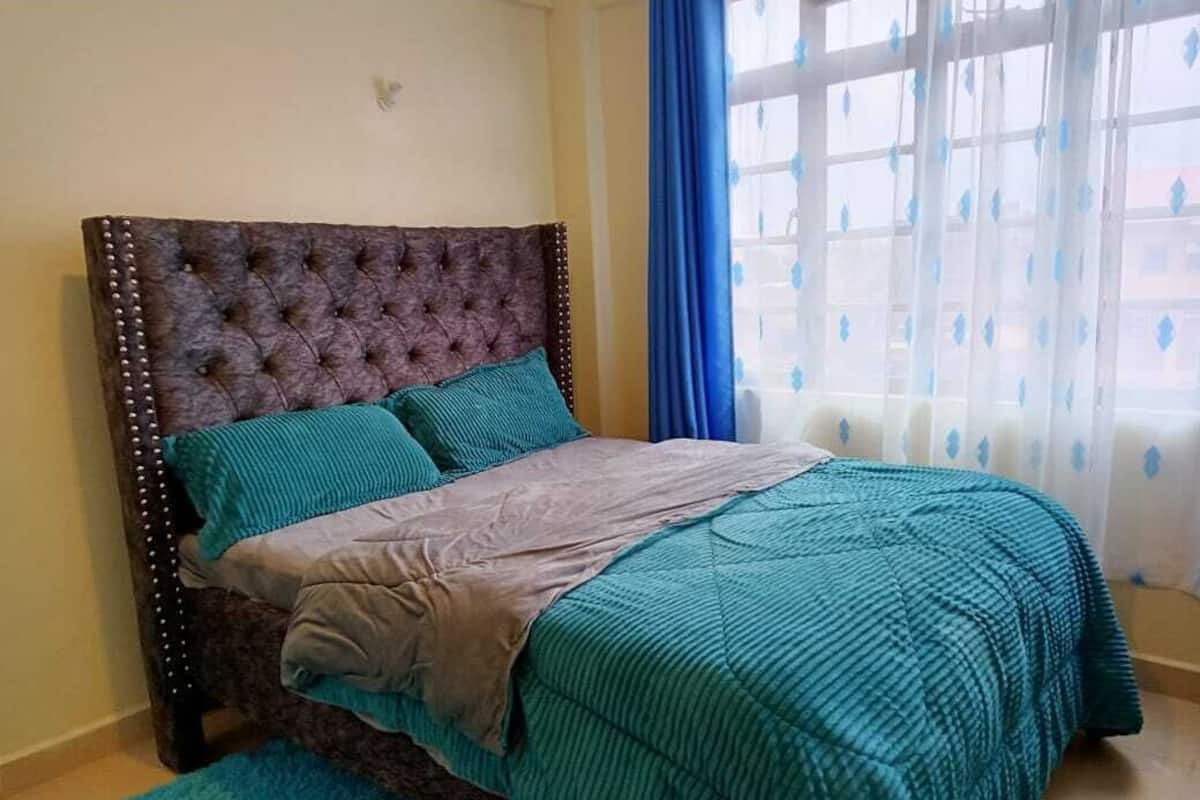 Une chambre à coucher avec une tête de lit capitonnée, une literie de couleur turquoise et une vue par une fenêtre ornée de rideaux bleus.