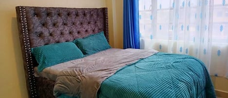A bedroom with a tufted headboard, teal bedding, and a view through a window with blue curtains.
